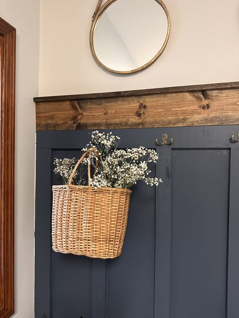 Vintage wicker basket filled with dried baby's breath hanging on a navy board-and-batten wall in a cottage-style mudroom.
