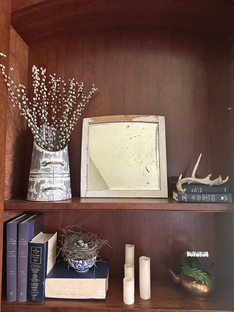 Vintage cottage shelf styled with an aged mirror, pussy willow branches, antique books, bird nest, antler, and cream-colored candles.