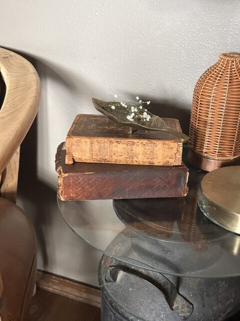 Stack of antique leather-bound books styled on a glass side table with a brass leaf dish and vintage-inspired decor.
