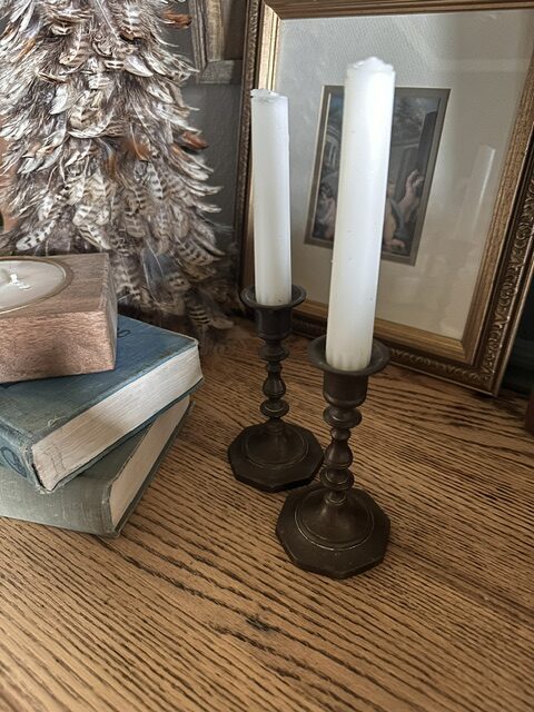 Vintage brass candlesticks with white taper candles displayed on stacked antique books beside a framed mirror and feather tree.