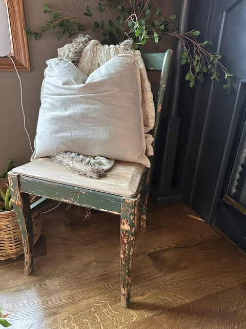 Vintage distressed green wooden chair styled with neutral linen pillows and a crocheted throw beside a black wood stove in a cozy cottage home.